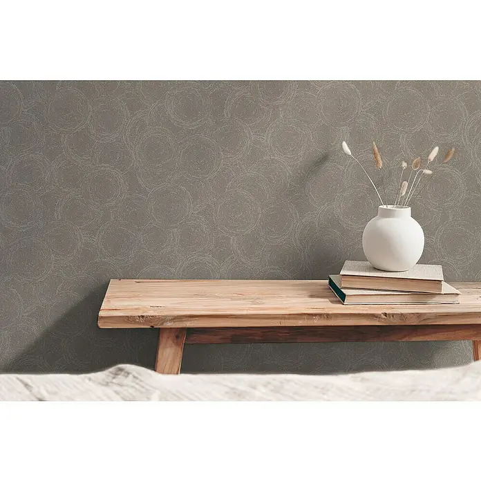 A.S. Création Hygge 3 Vliestapete Kreise - Braun-Beige-Grau, Grafisch, 10,05 x 0,53 m - Eine Nahaufnahme eines Raumes mit einer Holzbank, auf der Bücher und eine weiße Vase mit Pflanzen stehen, vor einer grauen Wand.