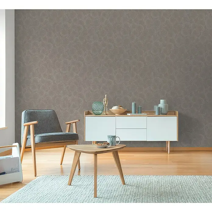 A.S. Création Hygge 3 Vliestapete Kreise - Braun-Beige-Grau, Grafisch, 10,05 x 0,53 m - Ein Wohnzimmer mit einem grauen Sessel, einem Couchtisch und einer weißen Kommode vor einer gemusterten Wand.