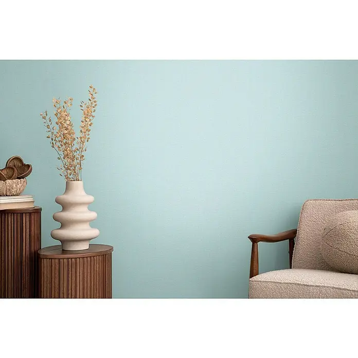 A.S. Création Cosmoliving by Cosmopolitan Vliestapete - Hellblau, Uni, 10,05 x 0,53 m - Dekorative Vase mit Trockenblumen auf einem Holzschrank, neben einem Sessel vor einer hellblauen Wand.