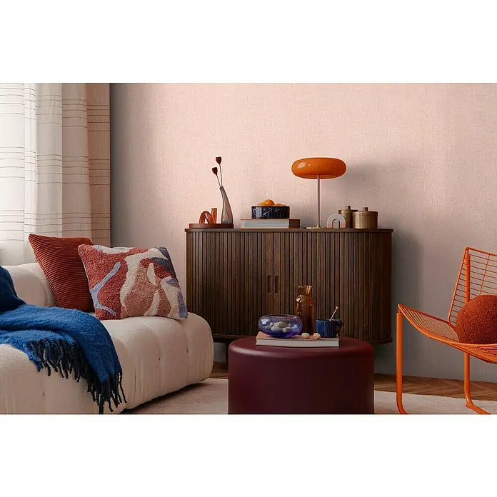 A.S. Création Cosmoliving by Cosmopolitan Vliestapete - Rosa meliert, Uni, 10,05 x 0,53 m - Wohnzimmer-Szene mit einem Lowboard aus Holz, einem Sessel mit Kissen und einer orangefarbenen Lampe, die eine gemütliche Atmosphäre schaffen.