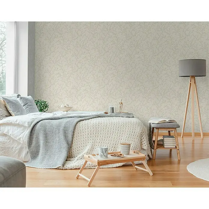A.S. Création Hygge 3 Vliestapete Ranken - Creme-Grau, Floral, 10,05 x 0,53 m - Schlafzimmer mit Bett, Nachttisch und Stehlampe mit grauem Schirm. Die Wände sind mit einer dezenten Tapete mit Blättermuster verziert.