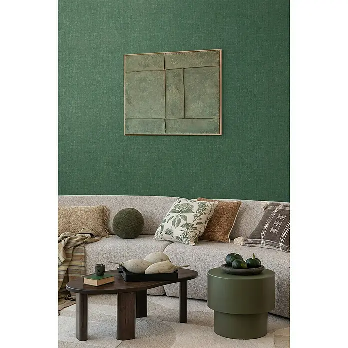 A.S. Création Cosmoliving by Cosmopolitan Vliestapete - Grün meliert, Uni, 10,05 x 0,53 m - Wohnzimmer mit olivgrünem Wandbild, beigem Sofa, Holztisch und dekorativen Elementen.