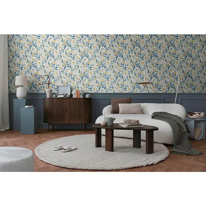 A.S. Création Cosmoliving by Cosmopolitan Vliestapete Blüte - Weiß-Blau-Gelb, Floral, 10,05 x 0,53 m - Wohnzimmer mit einem braunen Holzschrank, einem Sofa, einem Couchtisch und einem runden Teppich mit Hausschuhen darauf.
