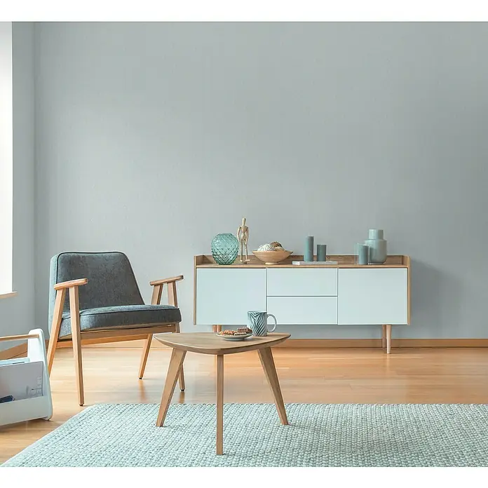 A.S. Création Hygge 3 Vliestapete - Hellblau, Uni, 10,05 x 0,53 m - Wohnzimmeraufnahme mit grauem Sessel, Beistelltisch mit Geschirr und einem Sideboard mit Dekorationsartikeln, die eine gemütliche Atmosphäre schaffen.