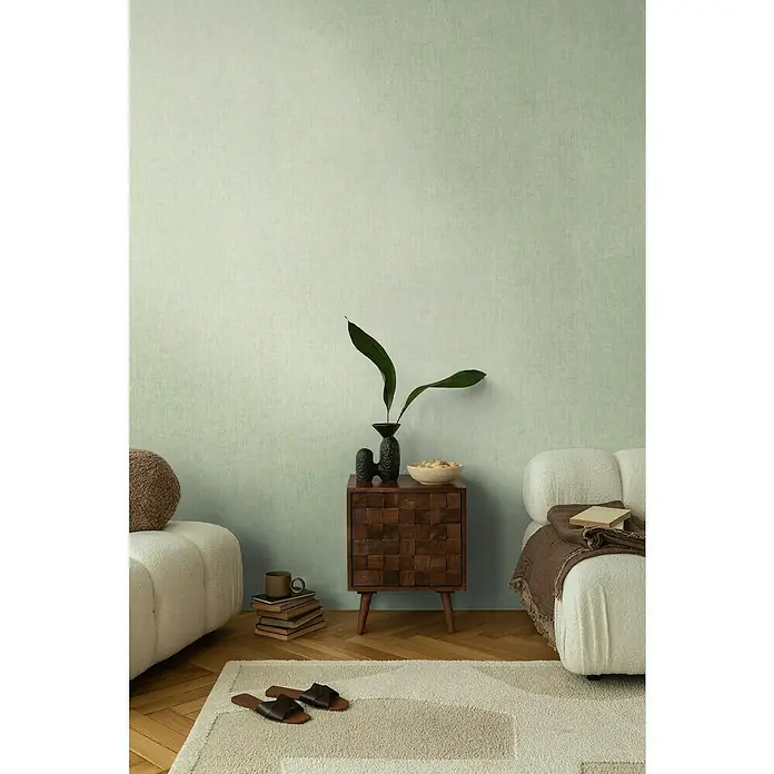 A.S. Création Cosmoliving by Cosmopolitan Vliestapete - Hellgrün, Uni, 10,05 x 0,53 m - Ein stilvolles Interieur mit einem Nachttisch aus dunklem Holz, dekoriert mit einer Vase und Pflanzen, neben einem Sofa und einem Teppich.