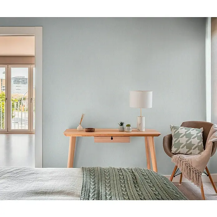 A.S. Création Hygge 3 Vliestapete - Hellblau, Uni, 10,05 x 0,53 m - Zimmeransicht mit einem Holztisch, Lampe, Stuhl mit Kissen und einer Tagesdecke auf einem Bett.