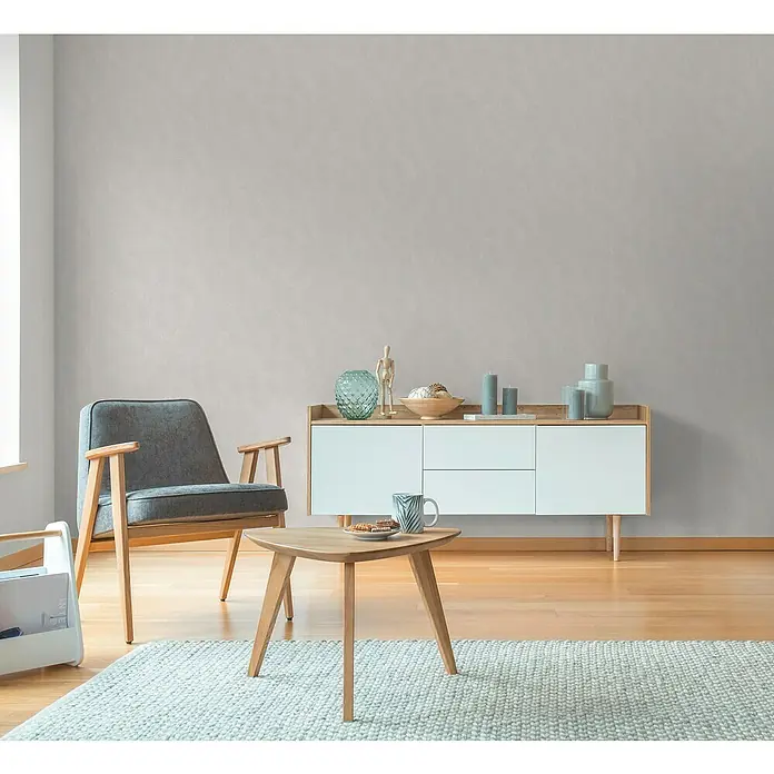 A.S. Création Hygge 3 Vliestapete Kreise - Creme-Weiß, Grafisch, 10,05 x 0,53 m - Wohnzimmer-Szene mit modernem grauen Sessel, hellem Teppich und niedrigem Sideboard. Die minimalistische Einrichtung strahlt Komfort und Stil aus.