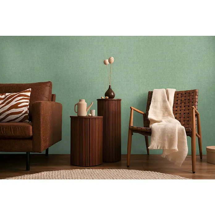 A.S. Création Cosmoliving by Cosmopolitan Vliestapete - Grün, Uni, 10,05 x 0,53 m - Zimmer-Interieur mit braunem Ledersofa, Rattansessel mit Wolldecke und gerippten Beistelltischen mit Dekor.