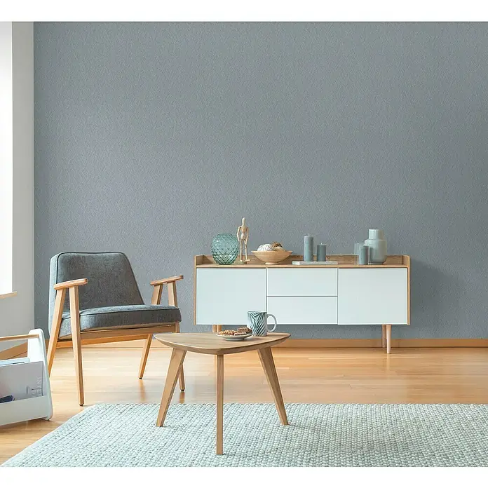 A.S. Création Hygge 3 Vliestapete - Grau-Blau, Uni, 10,05 x 0,53 m - Modernes Wohnzimmer mit einem grauen Sessel, einem stilvollen Sideboard und einem dreieckigen Couchtisch auf einem hellen Teppich.