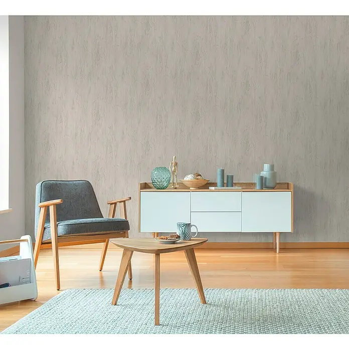 A.S. Création Hygge 3 Vliestapete Floral - Grau, Floral, 10,05 x 0,53 m - Wohnzimmer mit einem grauen Sessel, einem kleinen Tisch und einer weißen Kommode vor einer strukturierten Tapete.