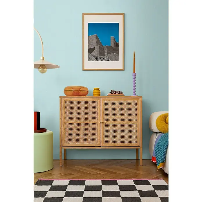 A.S. Création Cosmoliving by Cosmopolitan Vliestapete - Hellblau, Uni, 10,05 x 0,53 m - Einrichtungsszene mit einem Holzschrank mit Rattantüren, dekoriert mit Vasen und einem Kerzenständer, vor einer hellblauen Wand.