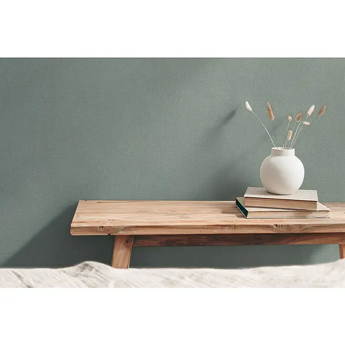A.S. Création Hygge 3 Vliestapete - Pastellgrün, Uni, 10,05 x 0,53 m - Stillleben mit einem rustikalen Holztisch, Büchern und einer Vase mit Trockenblumen vor einer schlichten Wand.