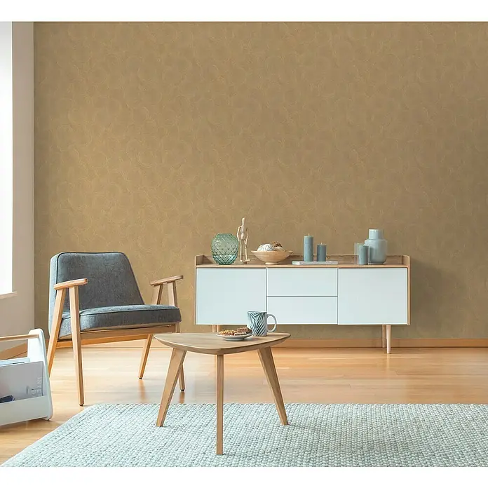 A.S. Création Hygge 3 Vliestapete Kreise - Braun-Gelb, Grafisch, 10,05 x 0,53 m - Wohnzimmer mit modernem Sessel, Tisch und Kommode vor einer goldenen Wandtapete.