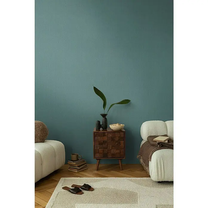 A.S. Création Cosmoliving by Cosmopolitan Vliestapete - Petrolgrün, Uni, 10,05 x 0,53 m - Stilvolles Interieur mit einem Nachttisch aus Holz, auf dem Vasen stehen, neben einem cremefarbenen Sofa auf einem gemusterten Teppich.