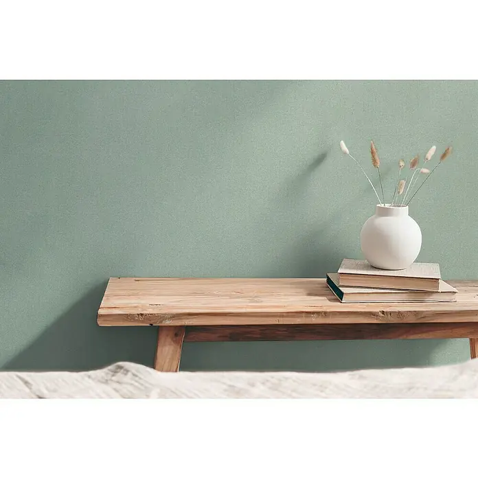 A.S. Création Hygge 3 Vliestapete - Mittelgrün, Uni, 10,05 x 0,53 m - Deko-Szene mit einer bauchigen, weißen Vase auf Büchern auf einer Holzbank vor einer mintfarbenen Wand, die ein minimalistisches und stilvolles Ambiente schafft.