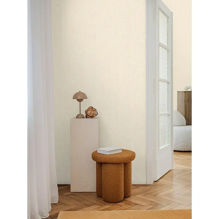 A.S. Création Cosmoliving by Cosmopolitan Vliestapete - Beige, Uni, 10,05 x 0,53 m - Eine Nahaufnahme eines stilvollen Raumes mit einem auffälligen, braunen Hocker und einem eleganten, beigen Sockel mit einer dekorativen Lampe.
