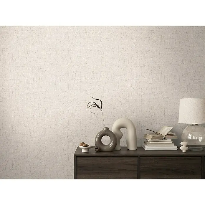 A.S. Création Cosmoliving by Cosmopolitan Vliestapete - Cremegrau, Uni, 10,05 x 0,53 m - Nahaufnahme eines Interieurs mit einer Kommode, Büchern, Vasen und einer Lampe vor einer beigen Wand mit Textur.