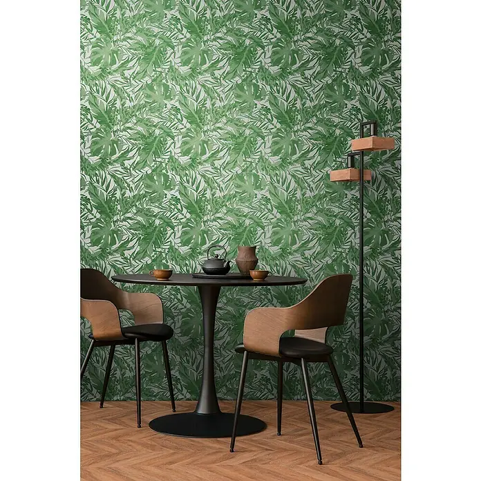 A.S. Création Cosmoliving by Cosmopolitan Vliestapete Blätter - Grün-Weiß, Floral, 10,05 x 0,53 m - Ein Essbereich mit einem schwarzen runden Tisch und Holzstühlen vor einer grünen Blättertapete, ergänzt durch eine moderne Stehlampe.