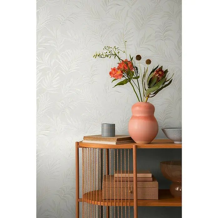 A.S. Création Cosmoliving by Cosmopolitan Vliestapete Ranken - Weiß-Creme, Floral, 10,05 x 0,53 m - Dekorative Nahaufnahme eines Sideboards mit Büchern, einer Vase mit Blumen und dekorativen Schalen vor einer blattgemusterten Tapete.