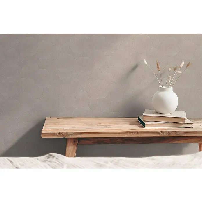 A.S. Création Hygge 3 Vliestapete Kreise - Beige-Grau, Grafisch, 10,05 x 0,53 m - Nahaufnahme einer weißen Vase mit getrockneten Gräsern auf einem Stapel Bücher, die auf einer Holzbank vor einer gemusterten Wand stehen.