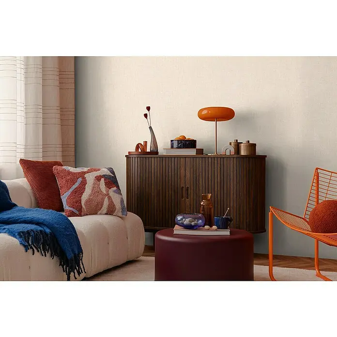 A.S. Création Cosmoliving by Cosmopolitan Vliestapete - Creme, Uni, 10,05 x 0,53 m - Wohnzimmer-Szene mit einem schicken Schrank, einer stilvollen Lampe, gemütlichen Kissen und einem orangefarbenen Stuhl, die eine warme Atmosphäre schaffen.