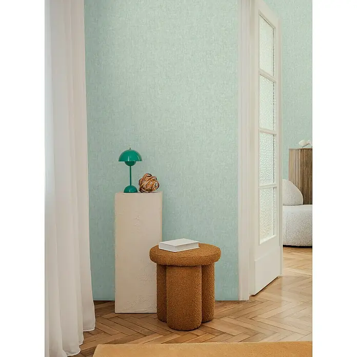 A.S. Création Cosmoliving by Cosmopolitan Vliestapete - Türkisgrün, Uni, 10,05 x 0,53 m - Zimmeransicht mit einem braunen, dreibeinigen Hocker mit Buch, einer grünen Lampe und einer weißen Säule vor einer mintgrünen Wand.