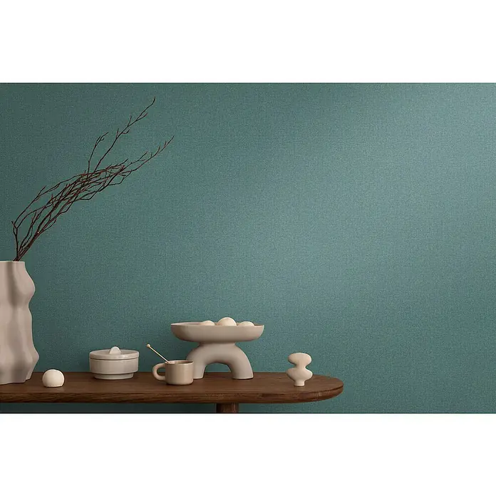 A.S. Création Cosmoliving by Cosmopolitan Vliestapete - Petrolgrün, Uni, 10,05 x 0,53 m - Dekoratives Arrangement mit einer Vase, einer Schale mit Kugeln, einer Dose, einer Tasse und einer Skulptur auf einem Holztisch vor blauem Hintergrund.