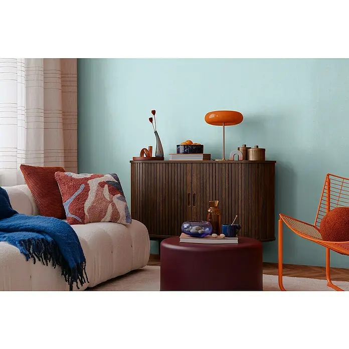A.S. Création Cosmoliving by Cosmopolitan Vliestapete - Hellblau, Uni, 10,05 x 0,53 m - Stilvolles Wohnzimmer mit einem Sideboard aus Holz, einer orangefarbenen Lampe und einem bequemen Sofa mit Kissen.
