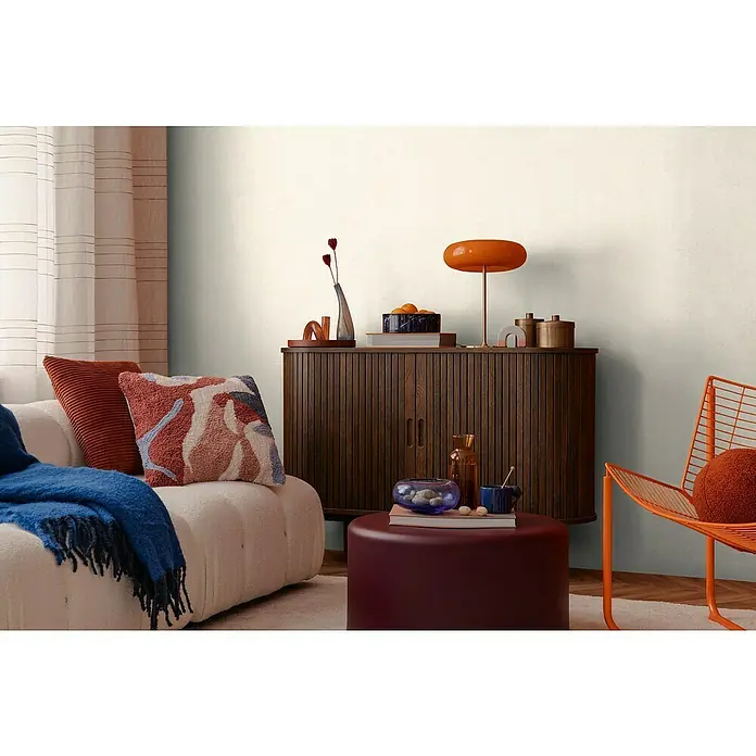 A.S. Création Cosmoliving by Cosmopolitan Vliestapete - Hellbeige, Uni, 10,05 x 0,53 m - Nahaufnahme von einem stilvollen Wohnzimmer mit einem Lowboard, einem Sofa mit Kissen, einem Hocker und einem orangefarbenen Stuhl.