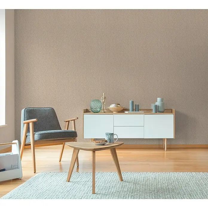 A.S. Création Hygge 3 Vliestapete - Braun, Uni, 10,05 x 0,53 m - Ein Wohnzimmer mit einem grauen Sessel, einem Couchtisch und einem Sideboard vor einer beigen Wand mit Muster.