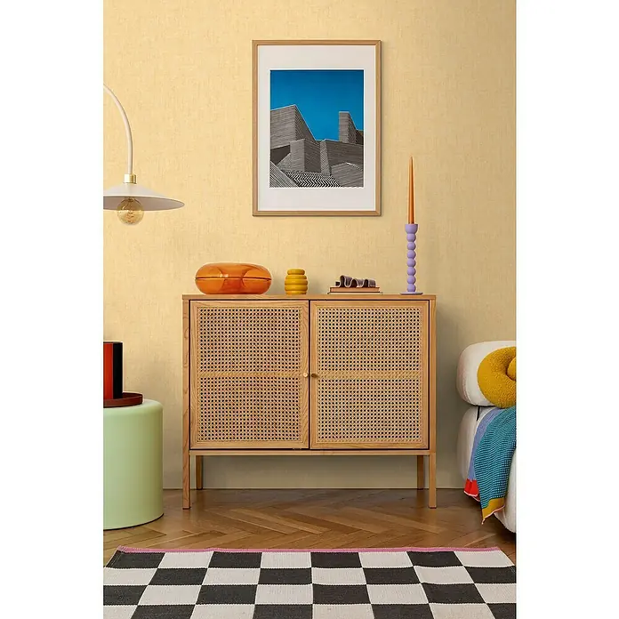 A.S. Création Cosmoliving by Cosmopolitan Vliestapete - Gelb, Uni, 10,05 x 0,53 m - Einrichtungsidee mit einem Holzschrank mit Rattantüren, dekoriert mit Vasen und einer Kerze, dazu ein modernes Gemälde und Schachbrettteppich.