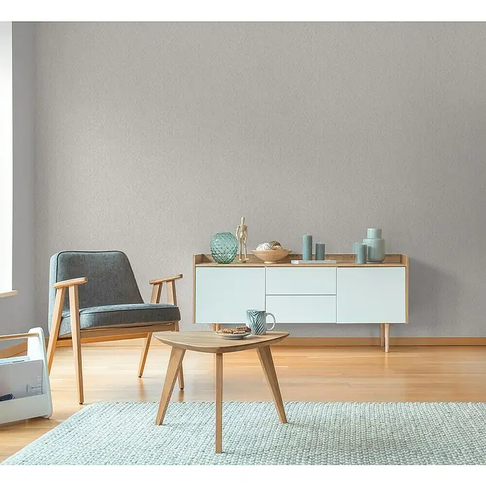 A.S. Création Hygge 3 Vliestapete - Hellgrau, Uni, 10,05 x 0,53 m - Helles Interieur mit grauem Sessel, Kommode mit Deko und einem Teppich. Fokus liegt auf der stilvollen Einrichtung des Raumes.