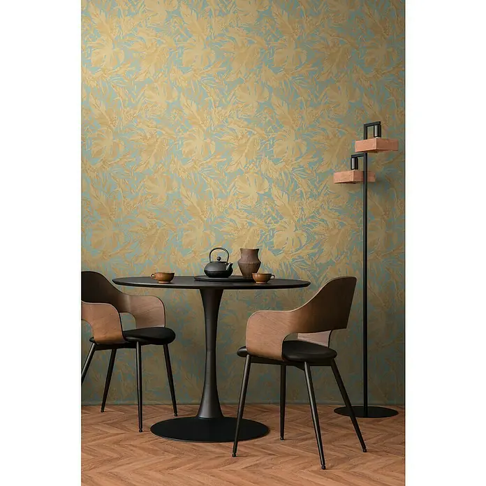 A.S. Création Cosmoliving by Cosmopolitan Vliestapete Blätter - Blau-Gold-Braun, Floral, 10,05 x 0,53 m - Ein Interieur mit einem schwarzen Esstisch und stilvollen Holzstühlen vor einer Wand mit Blattmuster. Eine Stehlampe rundet das Ambiente ab.