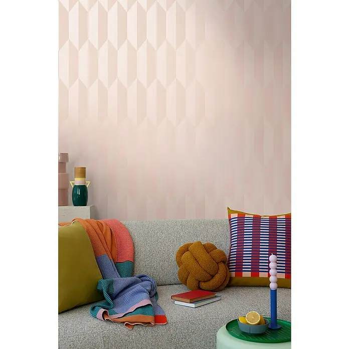 A.S. Création Cosmoliving by Cosmopolitan Vliestapete 3D-Grafik - Rosa-Metallic, Grafisch, 10,05 x 0,53 m - Das Bild zeigt ein helles Wohnzimmer mit einem grauen Sofa, farbenfrohen Kissen und Decken, sowie einer Dekoration auf einem Beistelltisch.