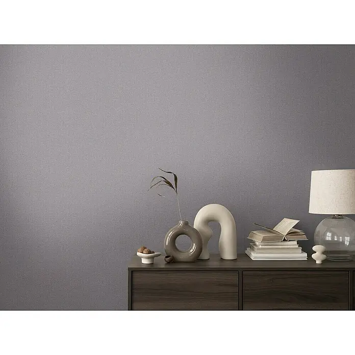A.S. Création Cosmoliving by Cosmopolitan Vliestapete - Dunkelgrau, Uni, 10,05 x 0,53 m - Nahaufnahme einer Kommode mit dekorativen Objekten wie einer Lampe, einer Vase und Büchern vor einer schlichten grauen Wand.