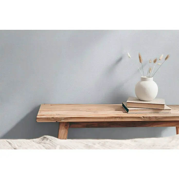 A.S. Création Hygge 3 Vliestapete - Pastelles Grau-Blau, Uni, 10,05 x 0,53 m - Schlichte Inneneinrichtung mit einer rustikalen Holzbank, Büchern und einer stilvollen weißen Vase mit Trockenblumen als Dekoration.