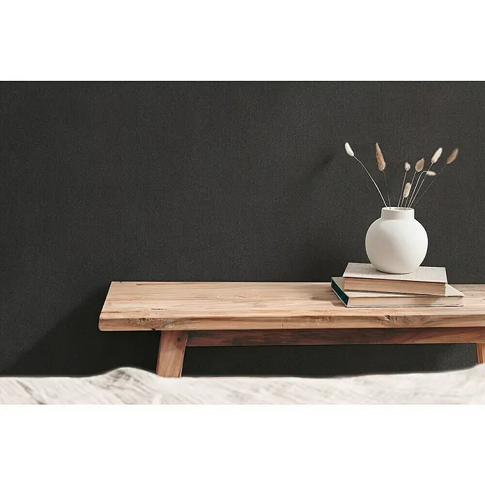A.S. Création Hygge 3 Vliestapete - Schwarz, Uni, 10,05 x 0,53 m - Schlichte Dekoration auf einem Holztisch mit einer weißen Vase und Büchern vor einer dunklen Wand. Natürliche und entspannte Atmosphäre.