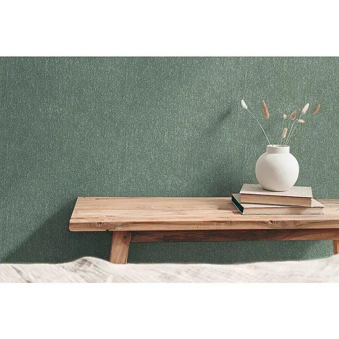 A.S. Création Hygge 3 Vliestapete Ranken - Grün, Floral, 10,05 x 0,53 m - Ein gemütliches Interieur mit einer Holzbank, Büchern und einer weißen Vase mit Gräsern vor einer strukturierten, grünen Wand.