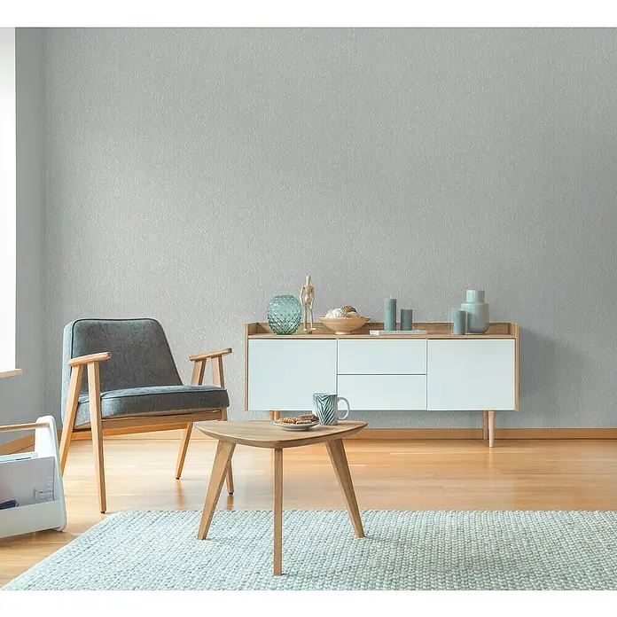 A.S. Création Hygge 3 Vliestapete Ranken - Grau, Floral, 10,05 x 0,53 m - Einladendes Wohnzimmer mit einem grauen Sessel, einem Holztisch und einem weißen Sideboard vor einer dezenten Tapete.