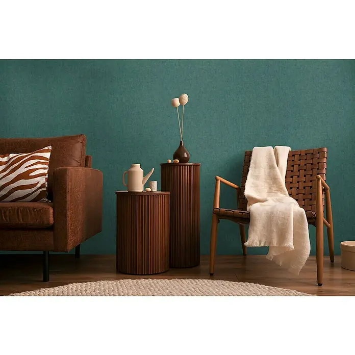 A.S. Création Cosmoliving by Cosmopolitan Vliestapete - Dunkelgrün, Uni, 10,05 x 0,53 m - Interieur mit braunem Ledersofa, Holzbeistelltischen und einem Stuhl mit cremefarbener Decke vor einer grünen Wand.