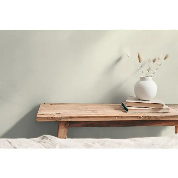 A.S. Création Hygge 3 Vliestapete - Hell-Creme, Uni, 10,05 x 0,53 m - Stimmungsvolles Interieur mit einer rustikalen Holzbank, Büchern und einer weißen Vase mit getrockneten Blumen vor einer schlichten Wand.