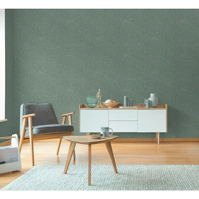 A.S. Création Hygge 3 Vliestapete Ranken - Grün, Floral, 10,05 x 0,53 m - Zimmer mit modernem Sessel, Tisch und Kommode vor einer grünen Wand.