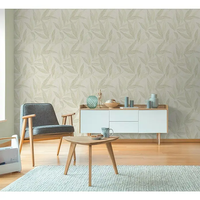 A.S. Création Hygge 3 Vliestapete Blätter - Creme-Grün-Silber, Floral, 10,05 x 0,53 m - Eine Inneneinrichtung mit einem grauen Sessel, Holztisch und einem weißen Sideboard vor einer Wand mit Blattmuster.