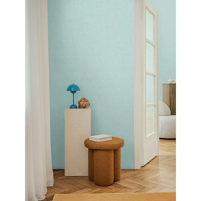 A.S. Création Cosmoliving by Cosmopolitan Vliestapete - Helles Blau, Uni, 10,05 x 0,53 m - Ein brauner Hocker aus Teddyplüsch mit einem Buch darauf vor einer hellblauen Wand. Er ist ein Hingucker in jedem Raum.