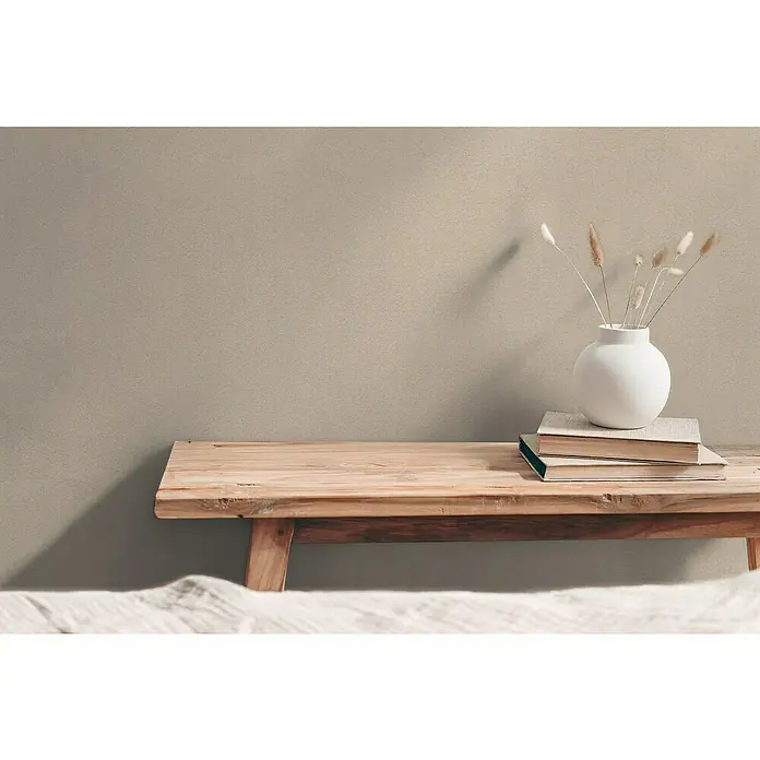 A.S. Création Hygge 3 Vliestapete - Hell-Braun, Uni, 10,05 x 0,53 m - Nahaufnahme einer minimalistischen Szene mit einer hölzernen Bank, Büchern und einer weißen Vase mit Trockenblumen vor einem schlichten Hintergrund.