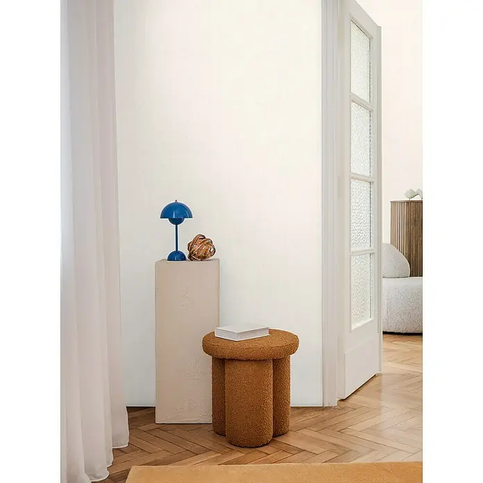 A.S. Création Cosmoliving by Cosmopolitan Vliestapete - Weiß, Uni, 10,05 x 0,53 m - Einrichtungsszene mit einem auffälligen, honigfarbenen Hocker mit drei Beinen und einem schicken blauen Design Lampe auf einem beigen Podest.