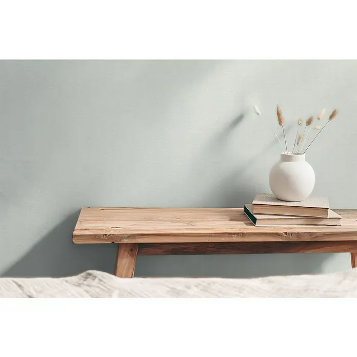 A.S. Création Hygge 3 Vliestapete - Offwhite, Uni, 10,05 x 0,53 m - Ein minimalistisches Stillleben mit einer weißen Kugelvase auf Büchern, die auf einer Holzbank vor einer hellgrünen Wand platziert sind.