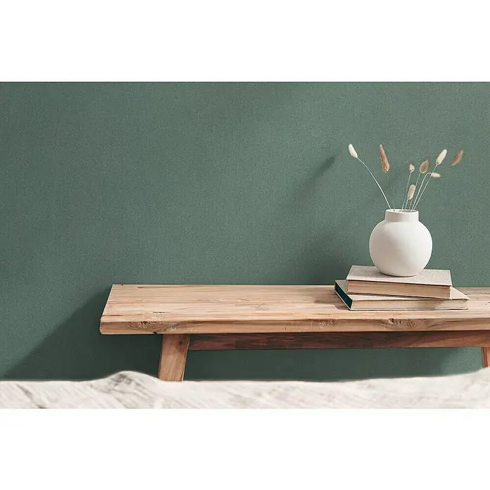 A.S. Création Hygge 3 Vliestapete - Grün II, Uni, 10,05 x 0,53 m - Stilleben mit Holzbank, Büchern und einer weißen Vase mit Gräsern vor einer grünen Wand, das eine ruhige und natürliche Atmosphäre vermittelt.
