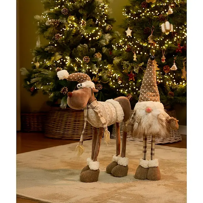Figura decorativa Vaala - L x An x Al: 25 x 16 x 69 cm, Textil - Una vista festiva con un reno y un gnomo de pie frente a árboles de Navidad brillantes, que irradian alegría navideña con su encanto.