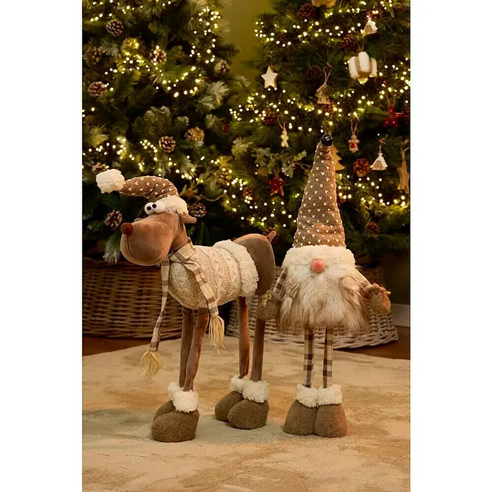 Figura decorativa Vaala - L x An x Al: 25 x 16 x 69 cm, Textil - Escena festiva con un reno de peluche con gorro y bufanda junto a un gnomo, con árboles de Navidad iluminados al fondo.